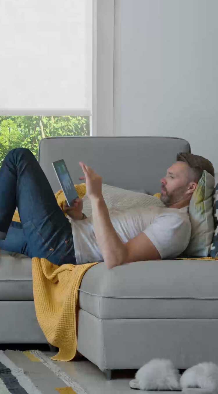 Man relaxing on couch with dog, playing on his iPad with roller shades filtering the outside light