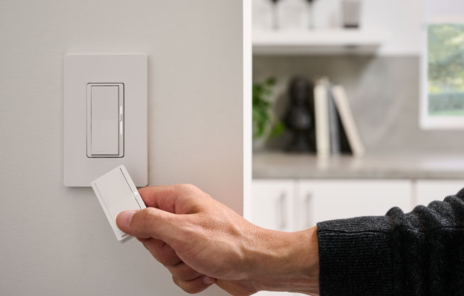 Male hand holding Pico Paddle Remote, pointing towards dimmer switch on wall